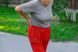 Older woman holding her lower back and hip while standing on a sidewalk, illustrating hip pain or osteoarthritis symptoms.