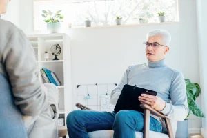 An older male pain psychologist with white hair and glasses holding a clipboard and listening attentively to a patient during a chronic pain behavioral health session at Augusta Pain Center